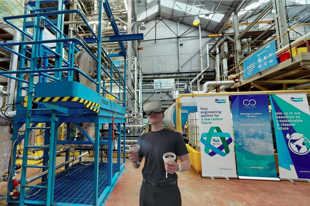 Image showing an industrial facility with pipes and machinery, a person wearing a VR headset, and display panels with Carbon Clean branding and CO₂-related messaging