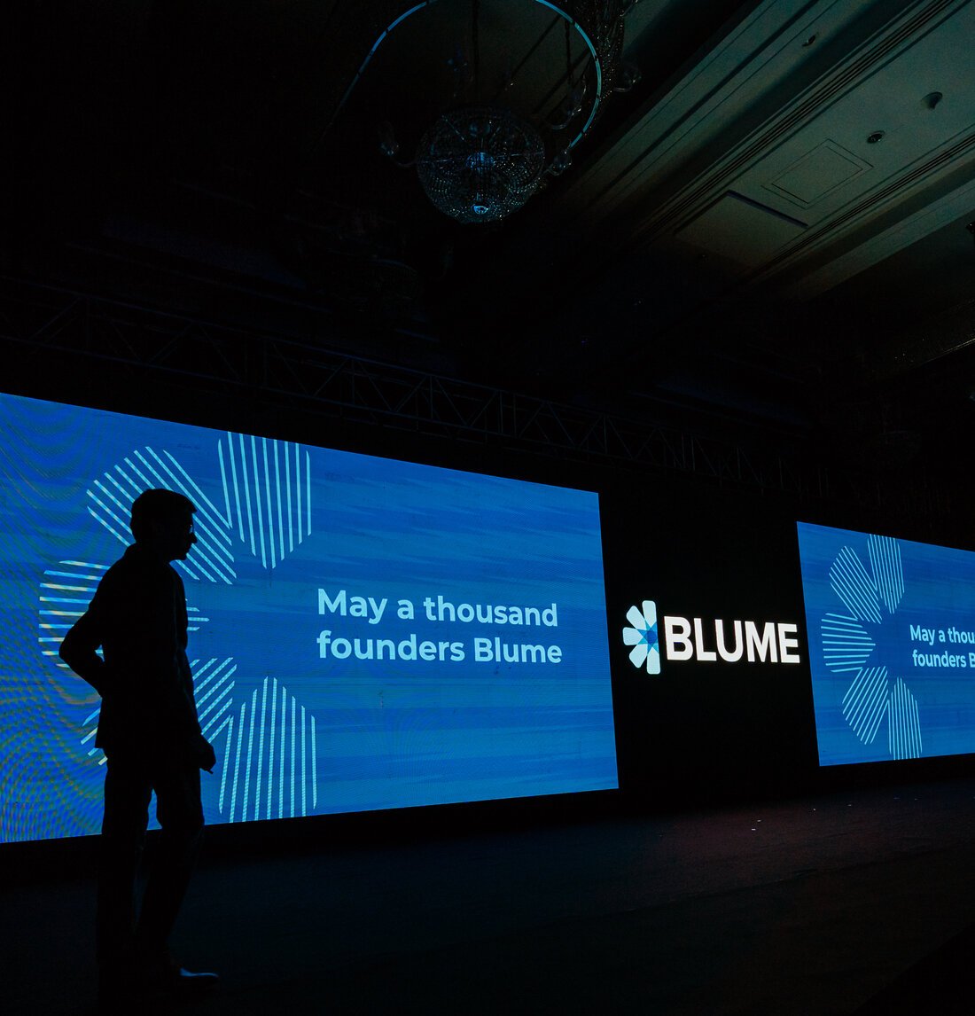 Image showing a stage with large screens displaying text May a thousand founders Blume and Blume branding with a person standing in silhouette